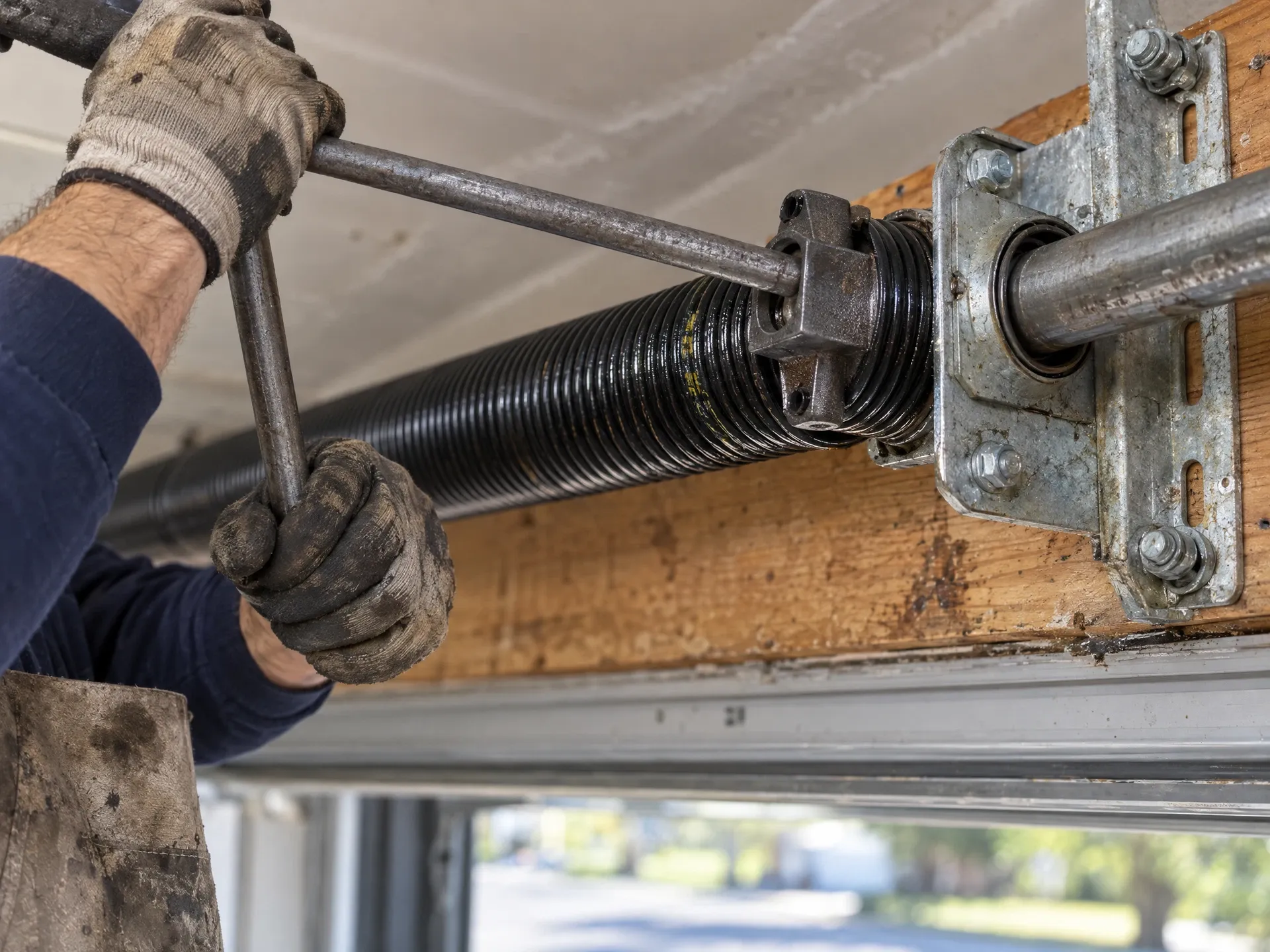 Gloved technician tensioning a new torsion spring on a residential garage door shaft with a winding bar